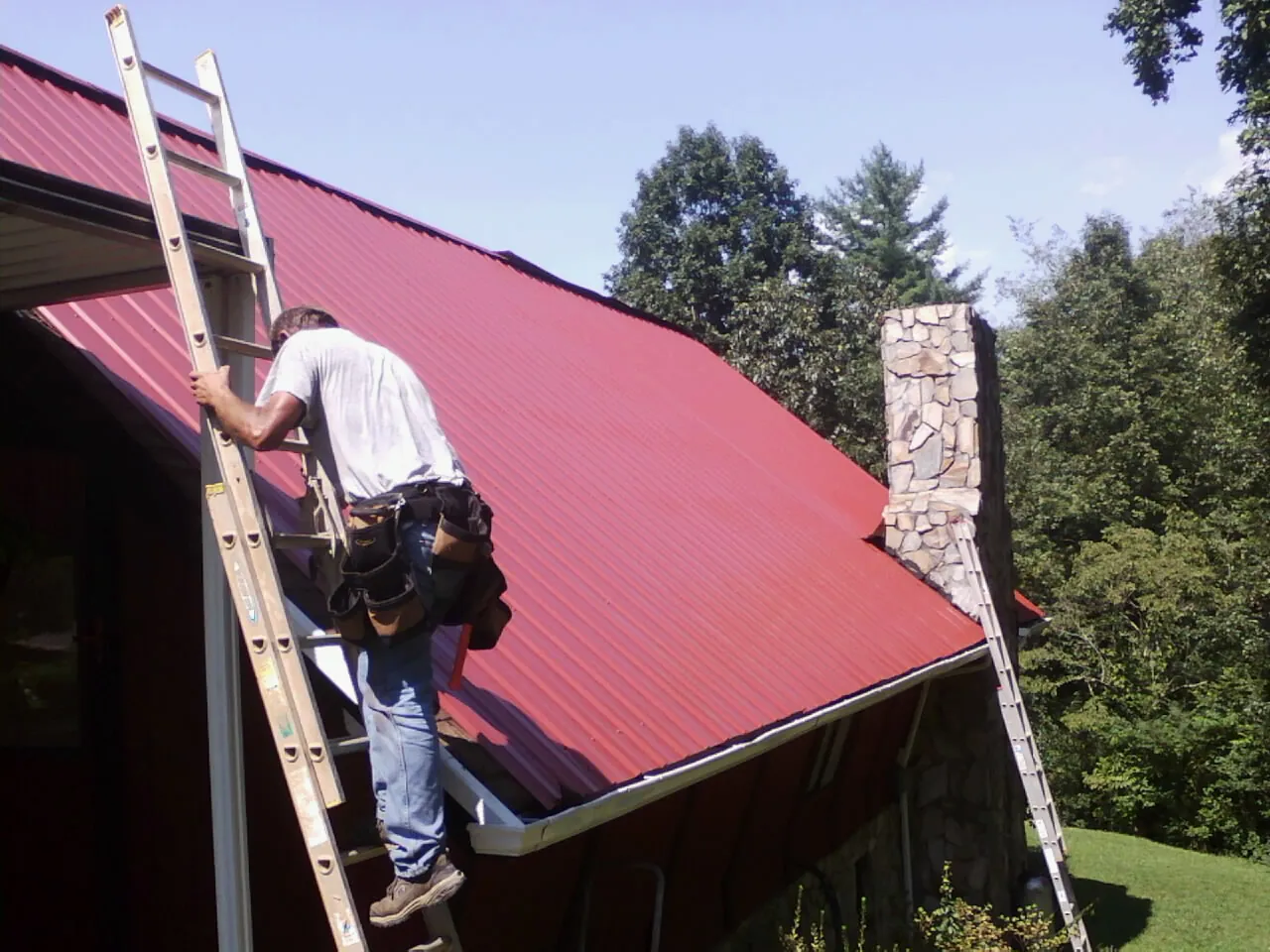 Roofer on a ladder beside a red metal roof and stone chimney — Soffit & Fascia Repair by Roofing Brothers in Sharon